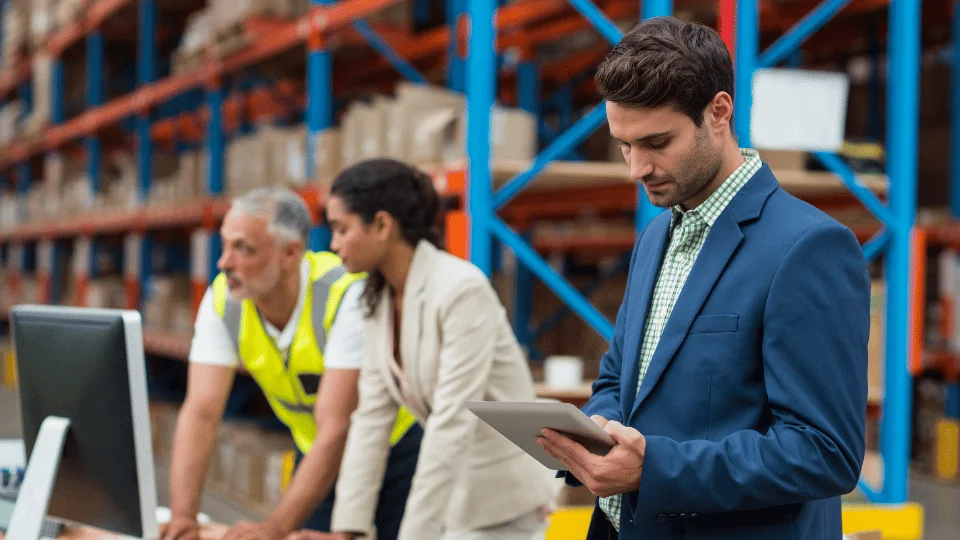 Freight Management Companies male manager looking at tablet while two warehouse workers are looking past computer while analyzing data