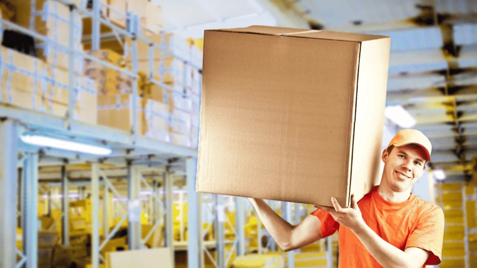 delivery man in orange holds up large square cardboard box on shoulders in warehouse